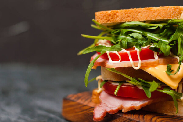 Huge tasty sandwich with ham or balyk, arugula, cheese, tomatoes and mayonnaise on a cutting board in a rustic style on gray background view from the side, copy space