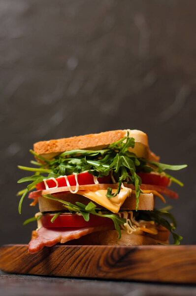 Огромный вкусный сэндвич с ветчиной или балыком, аругулой, сыром, помидорами и майонезом на разделочной доске в деревенском стиле на сером фоне сбоку, копировальное пространство
