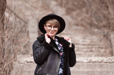 Siyah şapkalı, gözlüklü ve ceketli güzel, gülümseyen sarışın kadın sonbaharda şehir parkında merdivenlerde yürüyor.