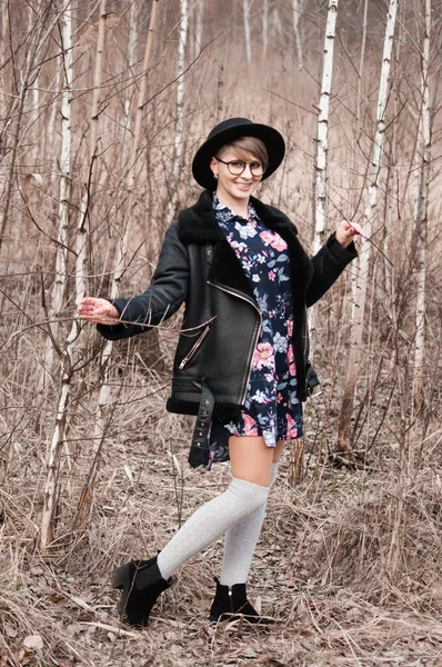 Sonbaharda ağaçların yanında poz veren şapkasız, gözlüklü ve ceketli güzel, mutlu sarışın kadın.