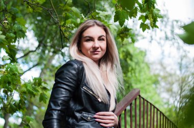 Baharda siyah deri ceketli güzel sarışın kız yeşil yaprakların, ağaçların arkasında güzel bir binanın yanında.