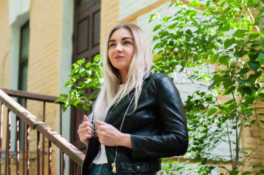 Baharda siyah deri ceketli güzel sarışın kız yeşil yaprakların, ağaçların arkasında güzel bir binanın yanında.