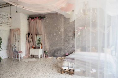 Bir fotoğraf stüdyosundaki eski bir odanın yan görüntüsü büyük aynalı beyaz bir ahşap makyaj masası, çiçekli vazolar, üzerinde pembe perdeden yapılmış bir elbise ve kitaplar ve çerçevelerle sarılı bir yatak.