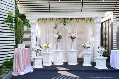 Çiçek ve perdelerle süslenmiş, kemer boyunca çiçekli vazolarla çevrili beyaz sütunlarla süslenmiş konukları karşılamak için güzel, beyaz, dikdörtgen bir düğün kemeri ve iki masa.