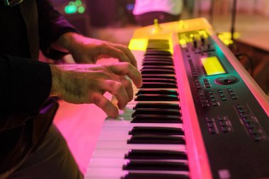 Genç müzisyenin elleri, sarı, pembe ve yeşil ışıklarla bir düğün ziyafeti salonunun sahnesindeki performansında elektrikli piyano tuşlarıyla çalar.