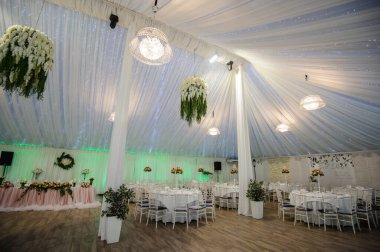 Bir düğün şölen salonunun içi: üzerinde beyaz masa örtüsü olan yuvarlak masalar ve üzerinde akşam yemeği için gerekli tüm malzemeler, çiçek buketi; beyaz perdeler, bej zemin, beyaz sandalyeler, pembe masa örtülü başkanlık masası