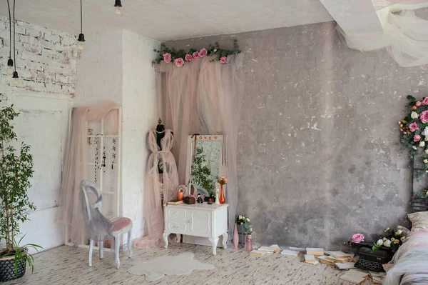 大きな鏡付きの白いヴィンテージの木製のドレッシングテーブル 花のある花瓶 椅子の横にピンクのカーテンからドレスを着た黒いマネキン 本に囲まれたベッドのある写真スタジオのヴィンテージルームの側面図 — ストック写真
