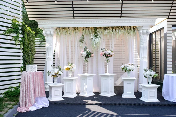 A beautiful white rectangular wedding arch for welcoming guests decoreted with flowers and curtains surrounded by white pillars with vases with flowers on them along the arch and two tables