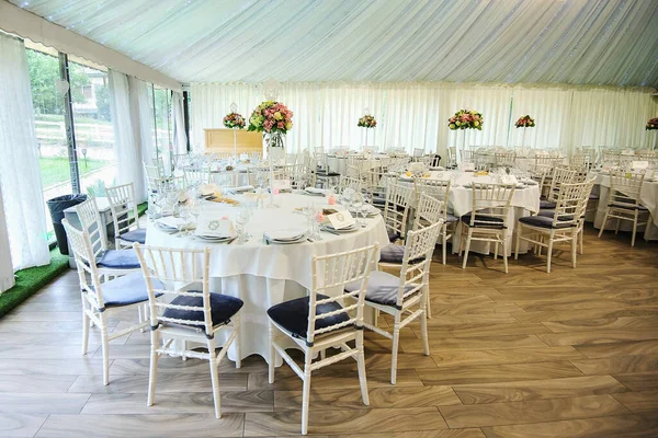 The interior of a wedding banquet hall: round tables with white tablecloth and all necessary supplies for dinner on them, bouquet of flowers; white curtains, beige floor, white chairs with blue pillows