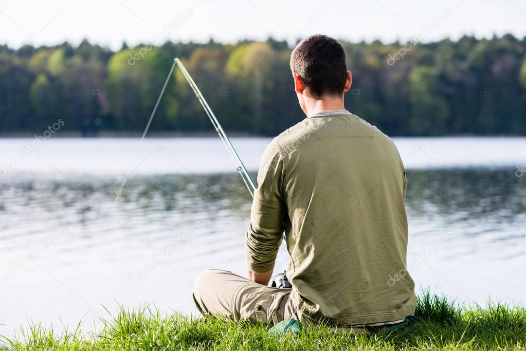 Angler sitting in grass at lake fishing with his rod