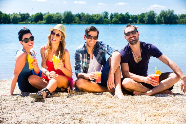 Quattro amici seduti sulla spiaggia del lago con cocktail — Foto Stock