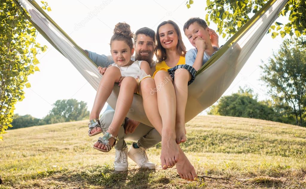 Family portrait with a beautiful mother of two playful children swinging in a hammock while looking at camera next to her husband outdoors in summer