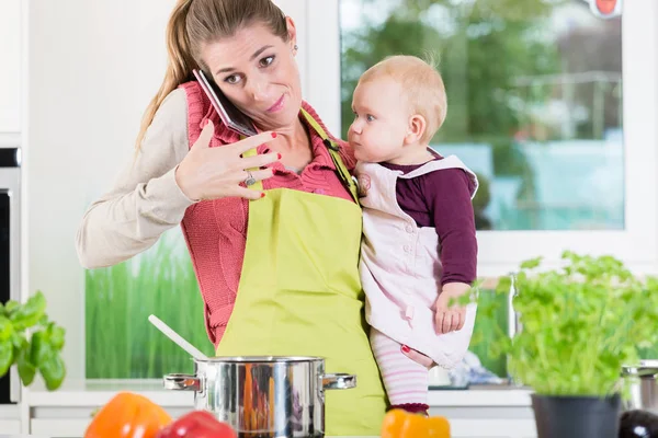 Madre teniendo conversación telefónica durante la cocción y sosteniendo al bebé — Foto de Stock
