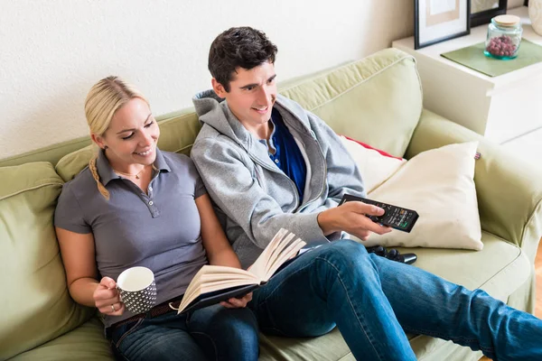 Mulher leitura livro ao lado de namorado assistindo TV — Fotografia de Stock