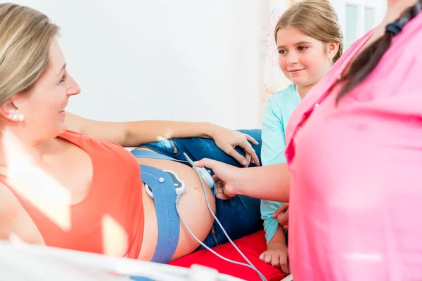 Молодая девушка смотрит акушерка прикрепляя CTG к беременному животу м — стоковое фото