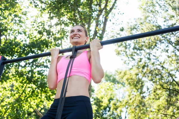 Cheerful young woman wearing pink sports bra while doing chin-up — Stock Photo, Image