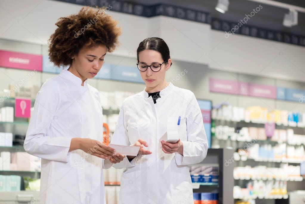 Two experienced female pharmacists wearing lab coats while analyzing together packages of new pharmaceutical drugs