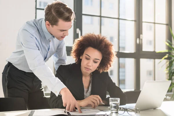 Supervisor checking business statistics and explaining results to colleague sitting at desk in office