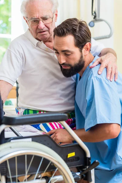 Elderly care nurse helping senior from bed to wheel chair — Stock Photo, Image
