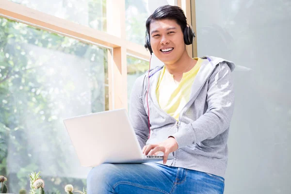 Hombre joven chino que usa ropa casual mientras usa una computadora portátil — Foto de Stock
