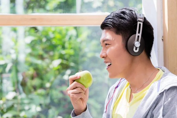 Jovem comendo uma maçã verde fresca enquanto ouve fones de ouvido — Fotografia de Stock
