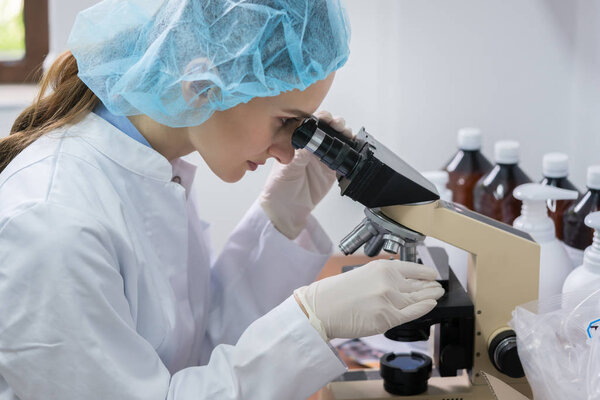 Side view of a female chemist wearing sterile safety equipment while analyzing sample under the microscope during work in the laboratory of a cosmetics factory