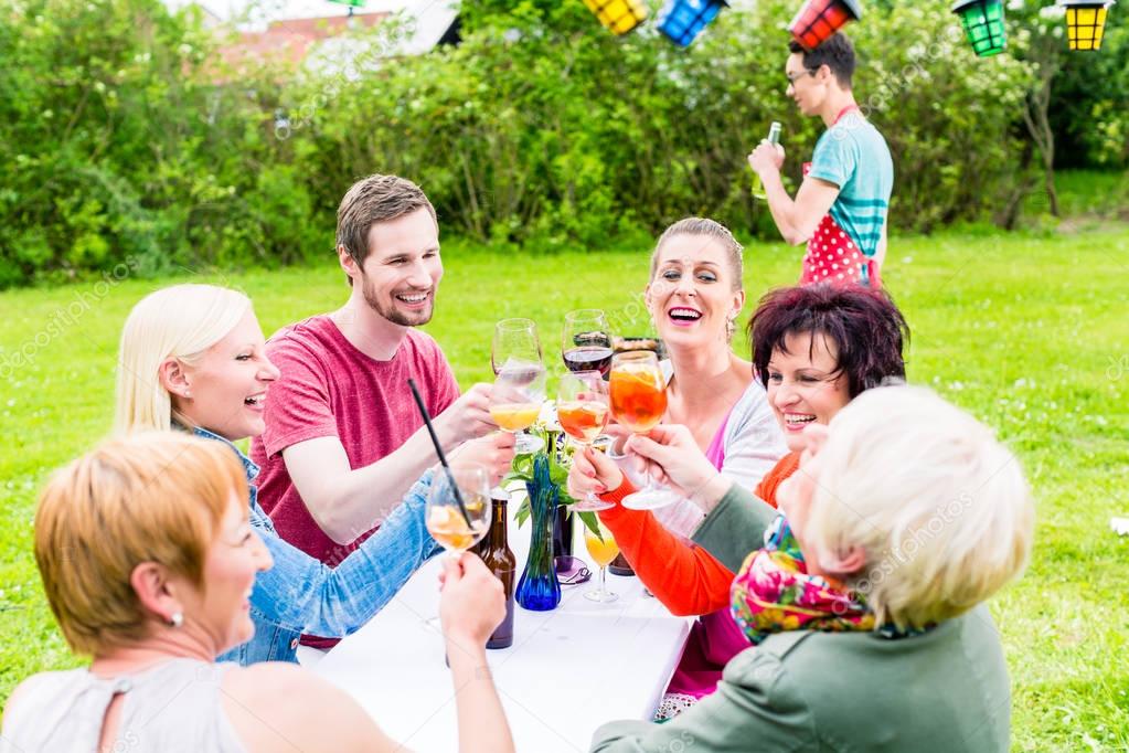 People toasting at party, in the background man at bbq grill with beer bottle in hand