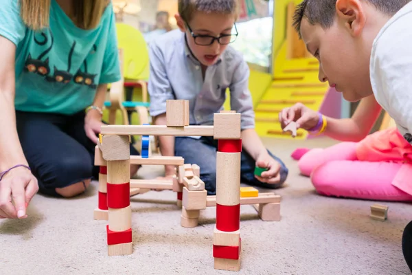 チャイルド自由遊び中に木製鉄道回路の構築と子どもたちを助ける専用の幼稚園の先生の低角度のビュー — ストック写真