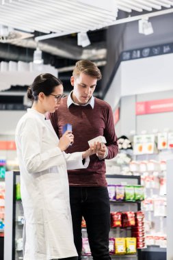 Experienced female pharmacist checking the indications and contraindications of a new medicine next to a young male customer in a modern pharmacy clipart