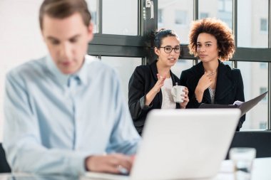 İki genç neşeli kadın çalışanların masa başında çalışan kendi meslektaşı yanındaki ofiste konuşmak