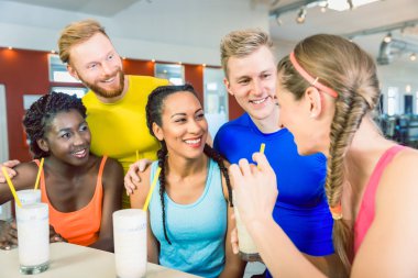 Çok etnik gruptan oluşan grup neşeli arkadaşıyla bir modern fitness kulübünde egzersiz önce beyaz protein karışımlarından yudumlarken gülüyor