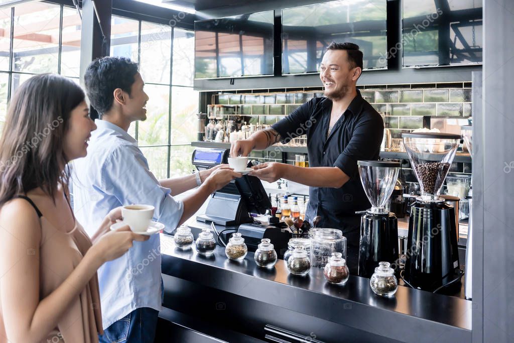 Young woman holding a white cup of coffee next to her partner served by a friendly barista in a modern coffee shop