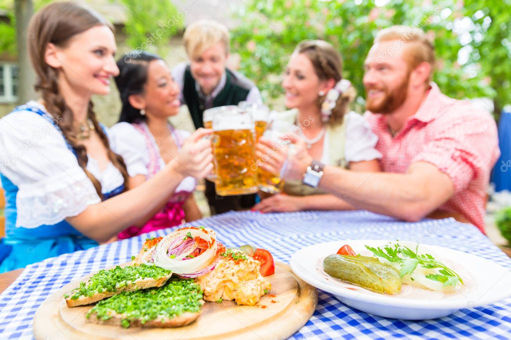 Five friends, women and men, sitting in beer garden clinking glasses having Bavarian appetizers on the table