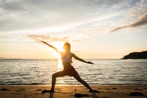 Silhueta de uma mulher apta praticando a pose de ioga guerreira ao pôr do sol — Fotografia de Stock