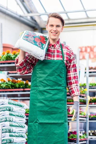 Pemuda ceria yang membawa sekantong tanah pot saat bekerja di toko bunga — Stok Foto