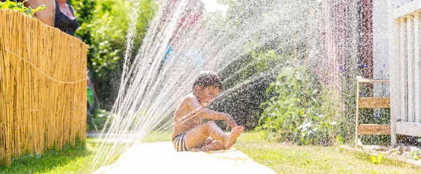 Niño enfriamiento con manguera de jardín, familia en el fondo —  Fotos de Stock