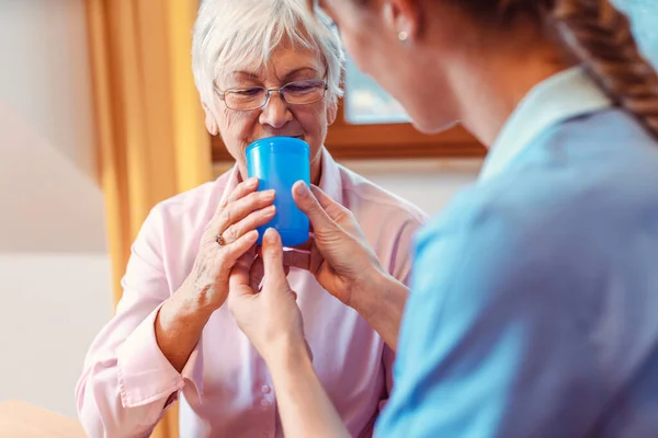 Смотритель помогает пожилой женщине пить, давая ей чашку воды — стоковое фото