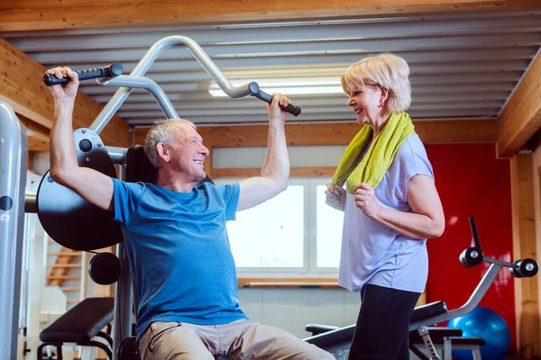 Homem sênior no ginásio com sua esposa em máquinas de exercícios — Fotografia de Stock
