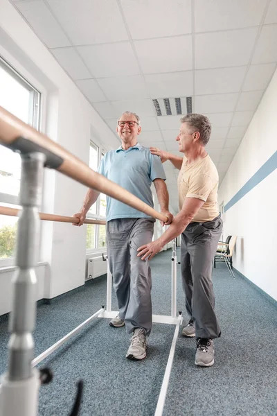 Fisioterapeuta ayudando a hombre mayor en rehabilitación caminando en los bares — Foto de Stock