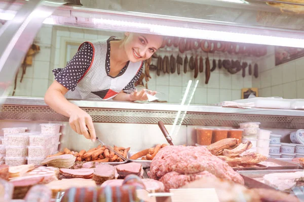 Vendedora en carnicería detrás de vitrina de cristal con carne y salchichas — Foto de Stock