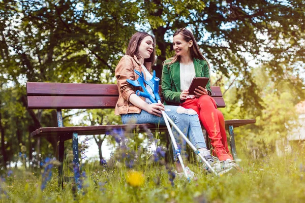 Dos amigos en un parque, uno con los pies rotos y muletas — Foto de Stock