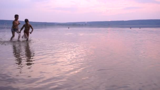 Två pojkar kör på floder stranden mot solnedgången — Stockvideo