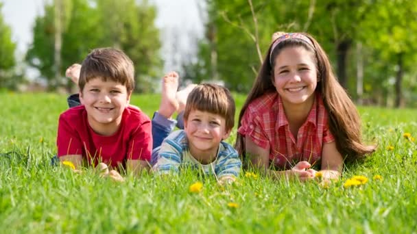 Trois enfants souriants couchés ensemble sur de l'herbe verte — Video