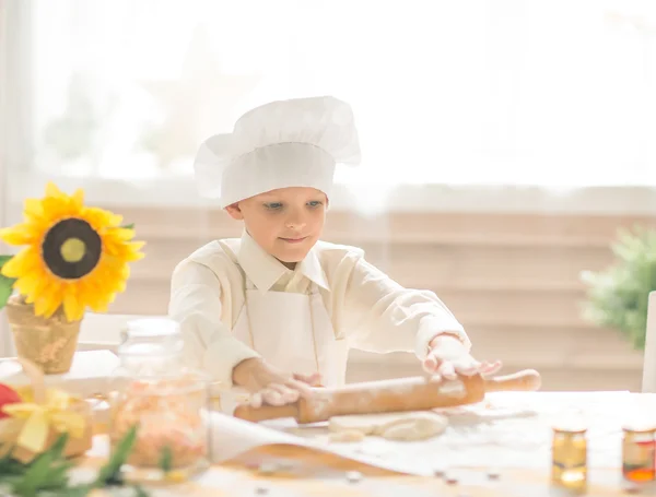 Маленький мальчик в форме повара выкатывает тесто на кухне — стоковое фото