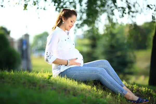 Grávida mãe senta-se na grama em um parque em um dia ensolarado . — Fotografia de Stock