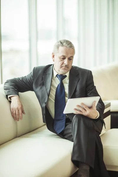 Hombre de negocios experimentado mirando la pantalla de una tableta digital sentada en un sofá en la oficina —  Fotos de Stock
