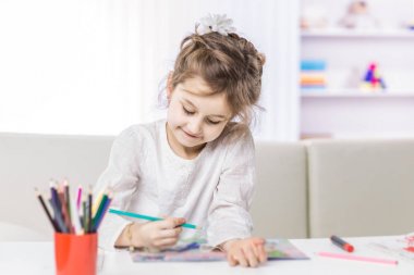 5 yaşındaki kız çocuk odasında masada oturan pastel boyalarla resim çiziyor.