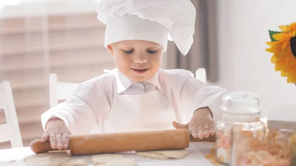 Uma criança pequena na forma de um cozinheiro rola a massa em uma mesa — Fotografia de Stock