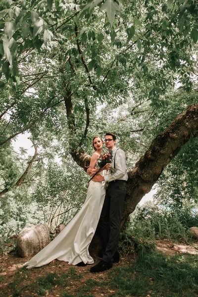 Ευτυχισμένη νύφη και γαμπρός στέκονται κοντά στο δέντρο . — Φωτογραφία Αρχείου