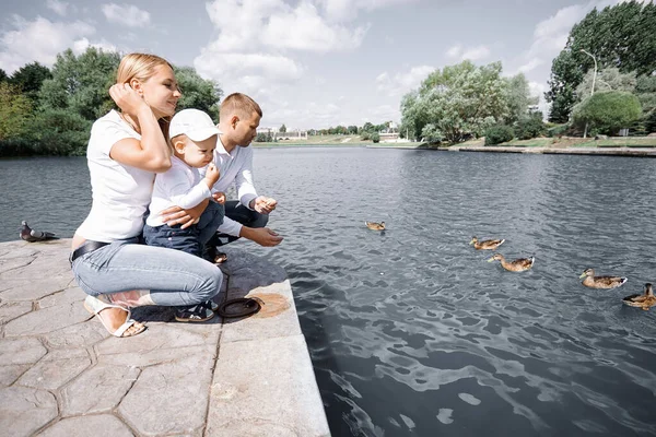 Familia joven alimenta a patos salvajes sentados en un muelle junto al lago — Foto de Stock